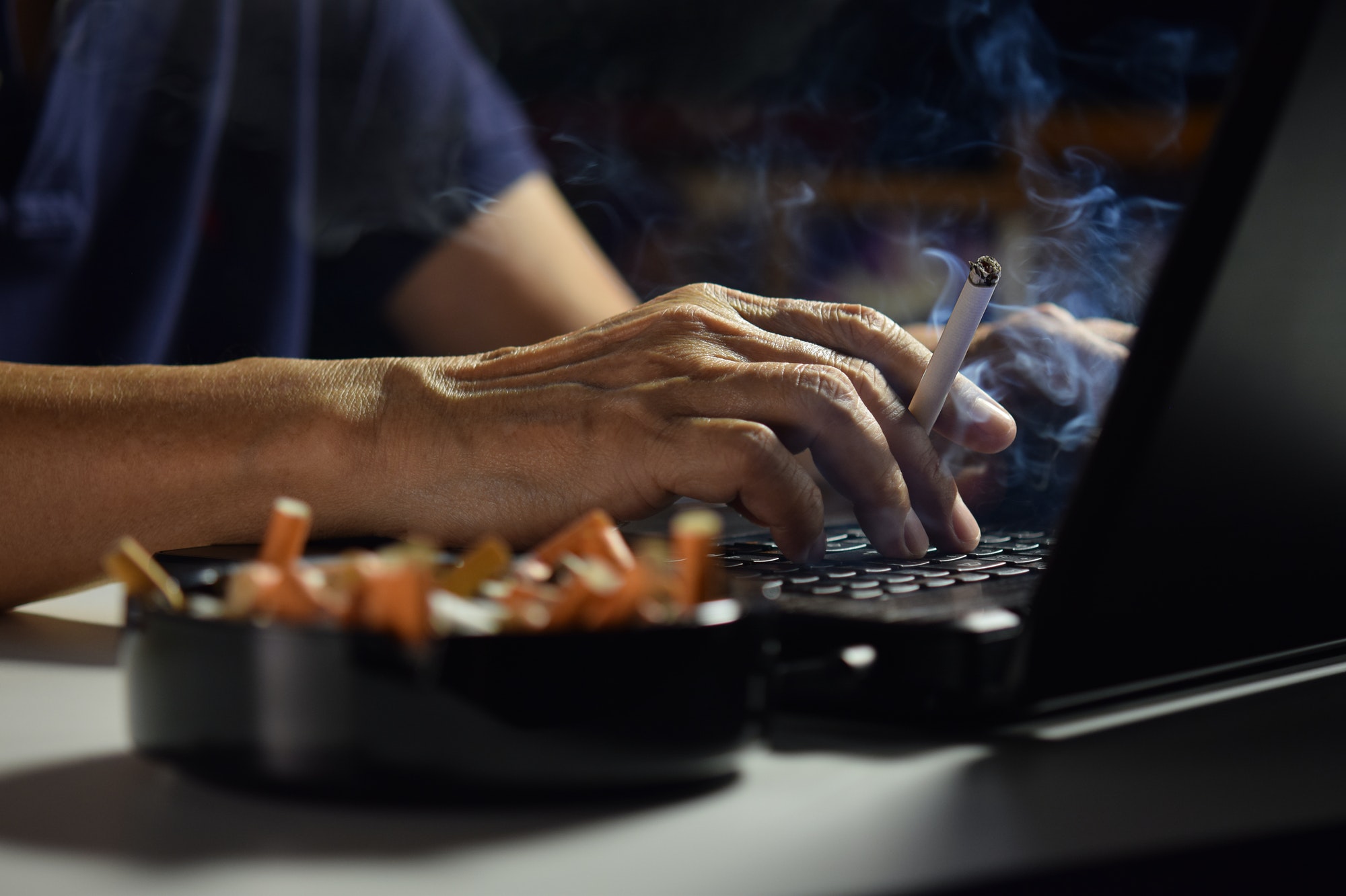 Man holding lit and smoking cigarette between fingers and working on a laptop computer